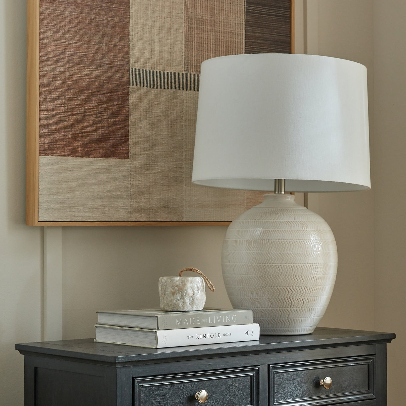 image of white ceramic table lamp with linen shade on a chest of drawers, next to a stack of books and in front of an art piece hanging on the wall