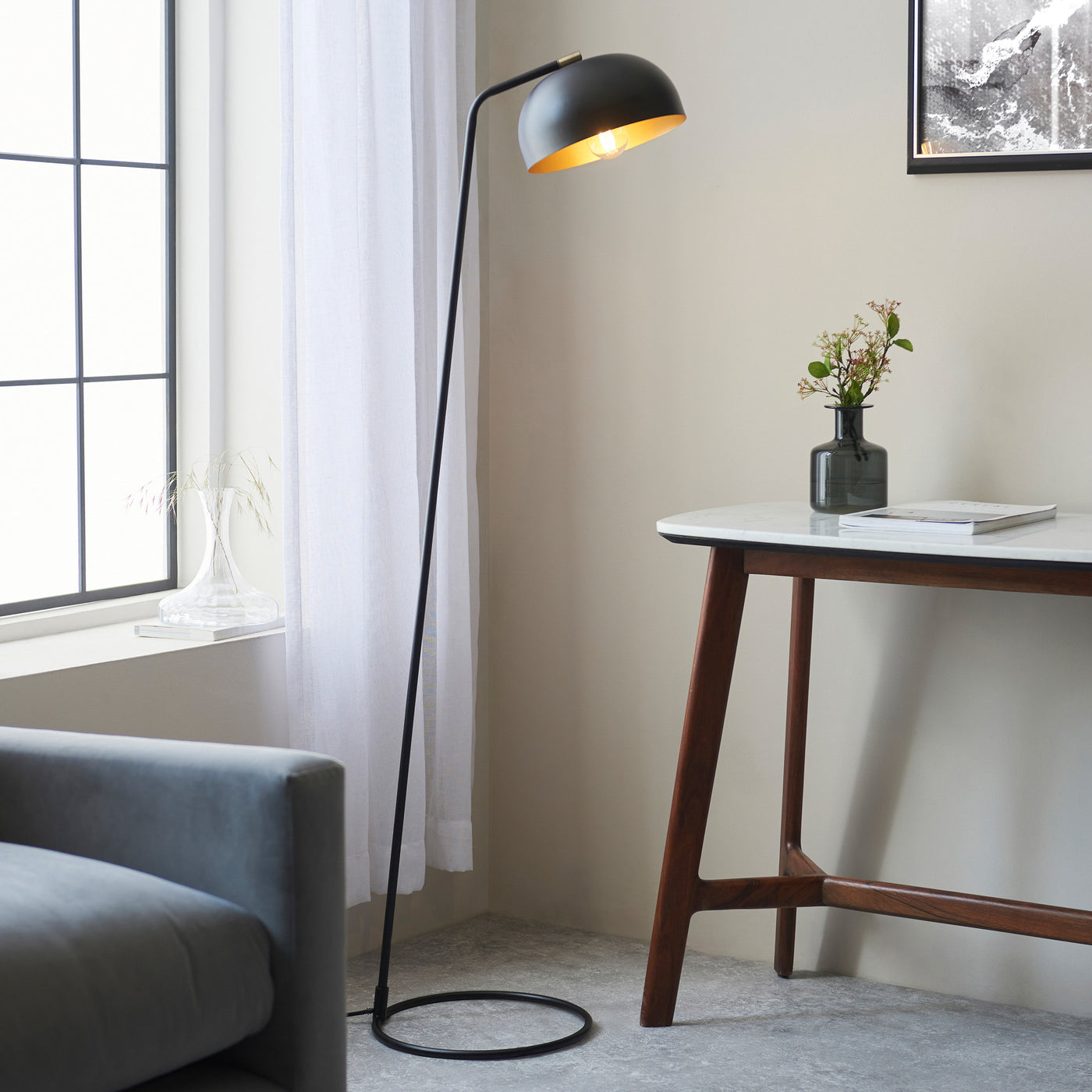 image of matt black thin floor lamp with curved head and domed shade next to grey sofa and modern brown console table