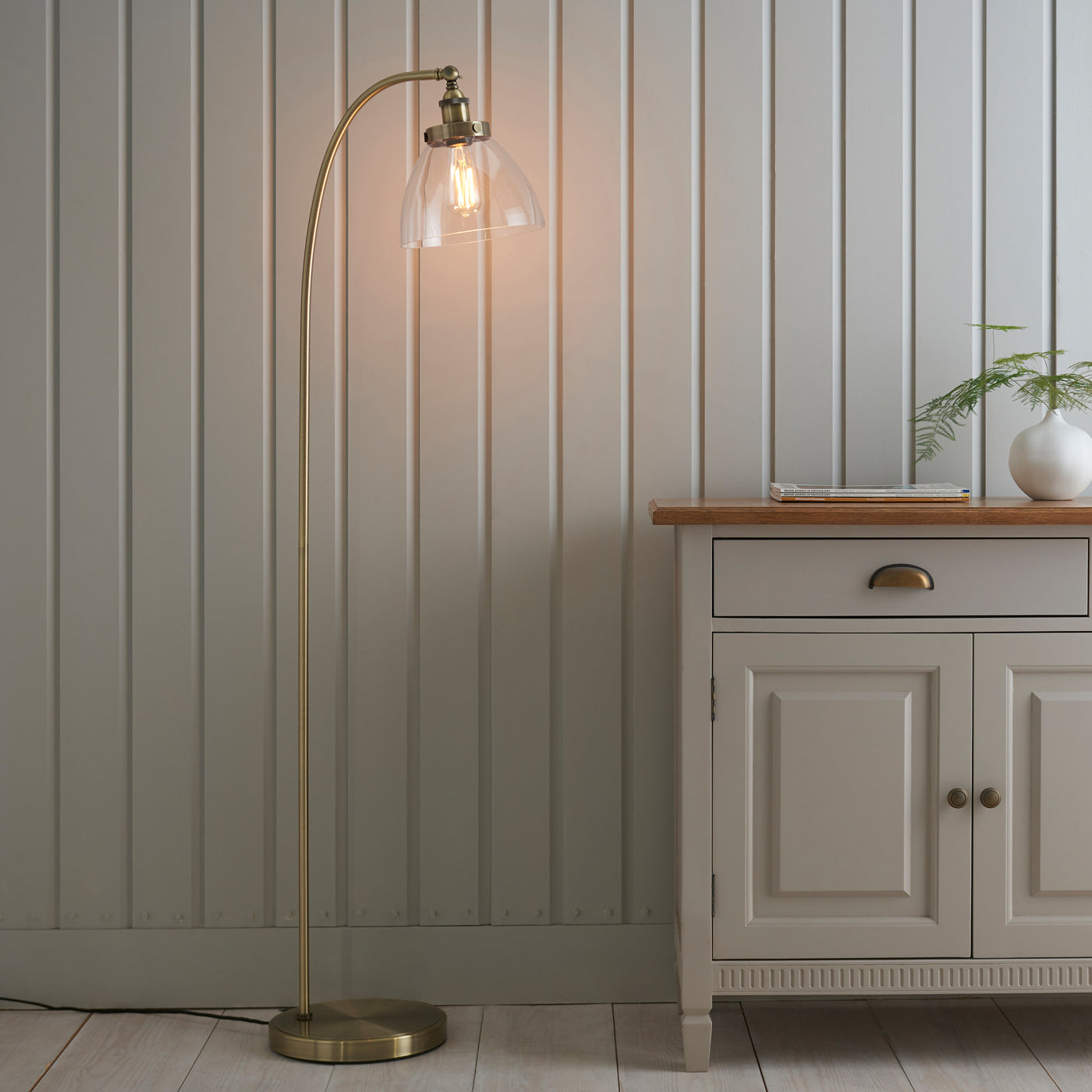 image of gently arched antique brass floor lamp with clear shade, stood in front of grey panelled wall next to grey sideboard, diffusing warm light