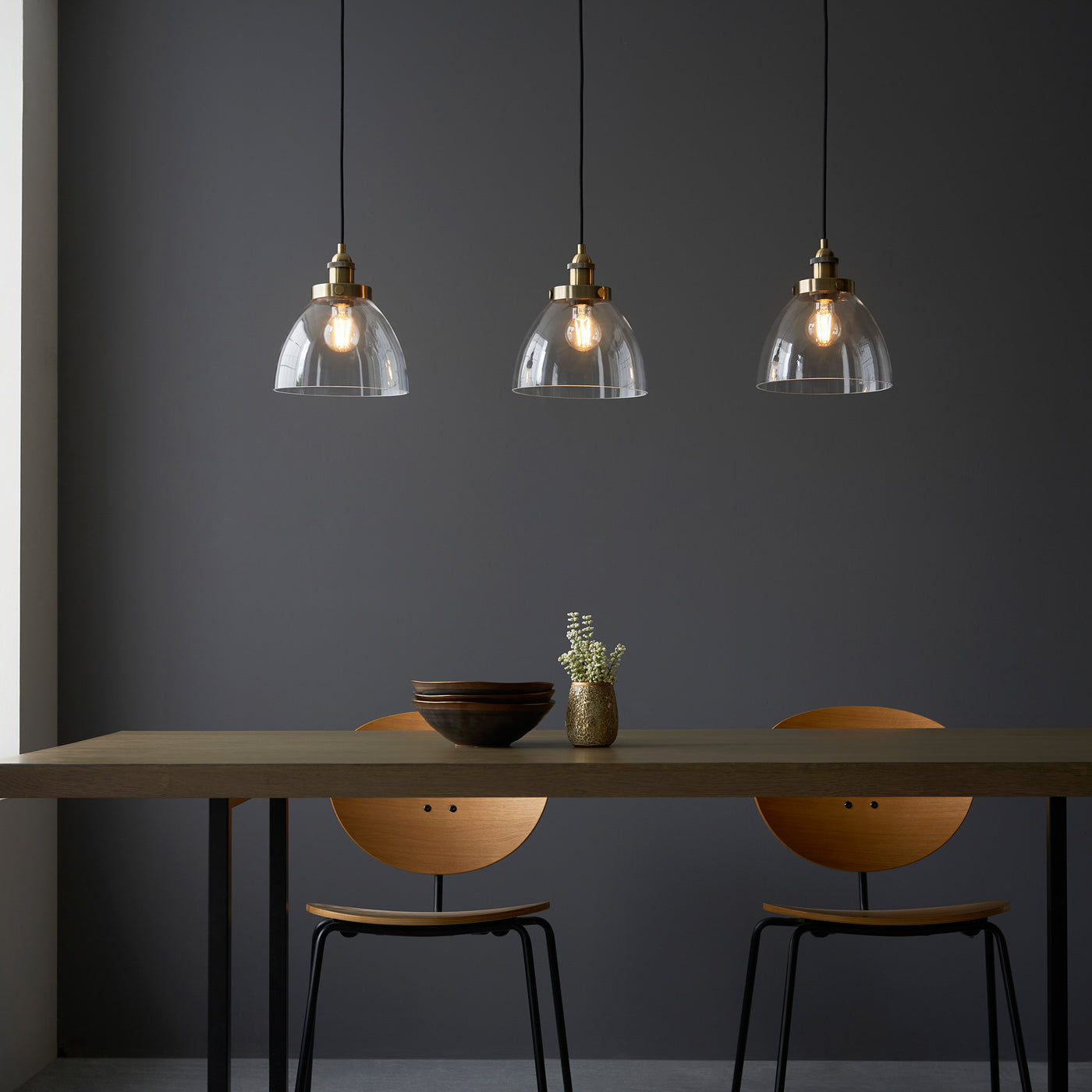 front on image of three clear glass light shades over warm white filament LEDs on a three-drop linear industrial antique brass light over dining table