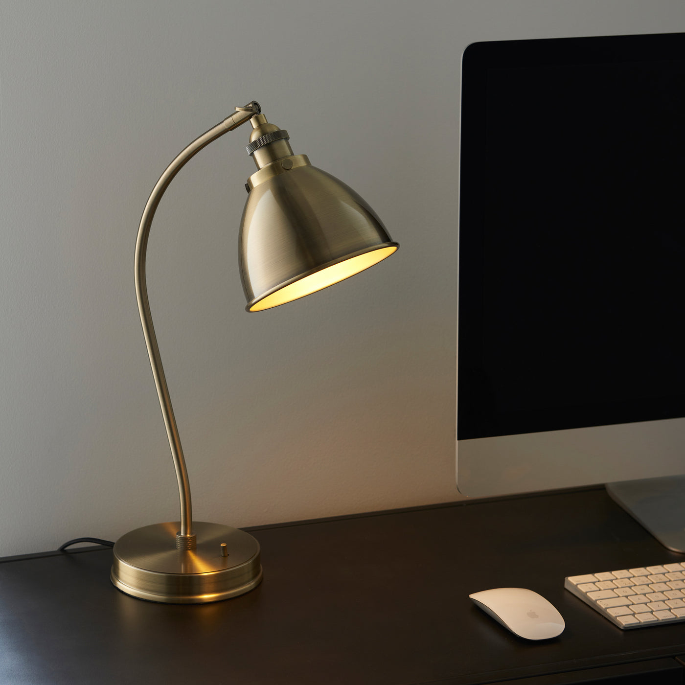 image of antique brass table lamp on a desk next to a computer, bulb switched on to emit a warm glow over the desk